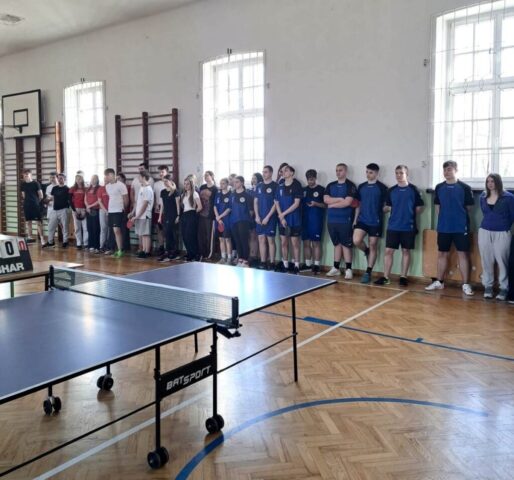 Uczestnicy wydarzenia sportowego stoją wzdłuż ściany sali gimnastycznej, obok stołu do tenisa stołowego przygotowanego do rozgrywek.
