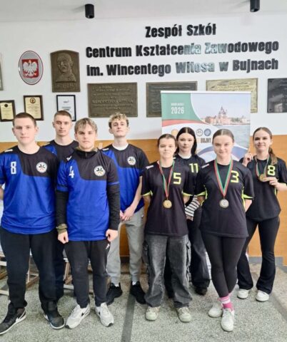Grupa uczniów w niebieskich i czarnych strojach sportowych pozuje w korytarzu szkoły; część z nich ma na szyjach medale.