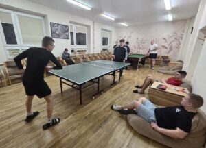 Kilka młodych osób wewnątrz świetlicy. Pośrodku rozłożony stół do ping-ponga i dwóch zawodników. W tle inne osoby przy stole do bilarda i 3 osoby siedzące po bokach. 