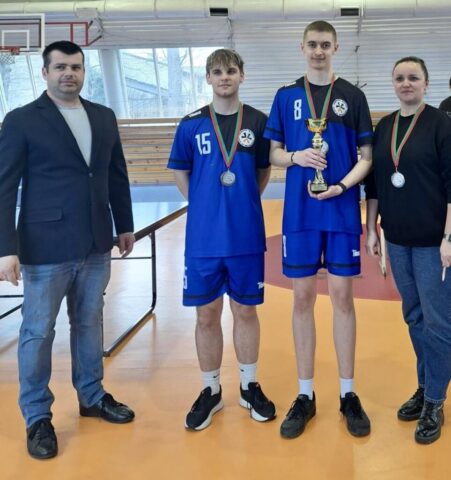 Dwaj uczniowie z medalami i pucharem pozują do zdjęcia wraz z opiekunami w sali gimnastycznej.