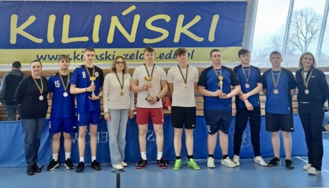 Grupa uczestników zawodów sportowych pozuje z medalami i pucharami na tle szkolnego baneru.