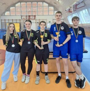 Pięciu uczestników zawodów sportowych pozuje z medalami i pucharami na sali gimnastycznej.