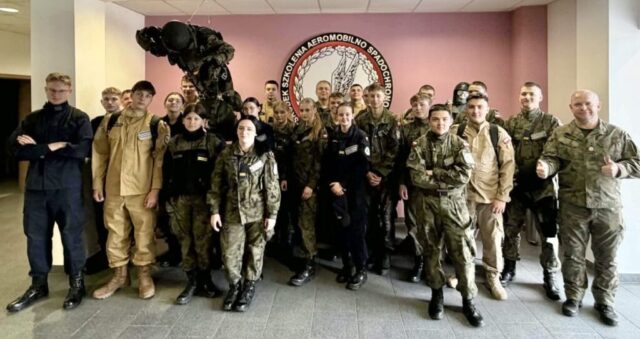 Grupa osób, młodzież, kadeci oddziału OPW wraz z opiekunem, w różnych mundurach, stoi w pomieszczeniu przed dużym emblematem na ścianie.