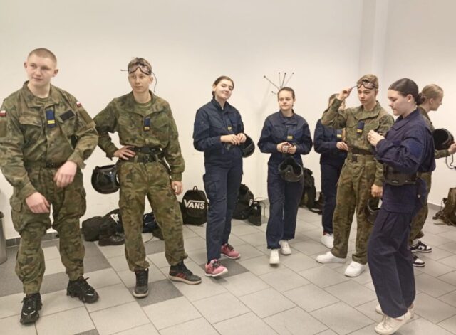 Grupa 8 kadetów klas OPW w mundurach i kombinezonach stoi w pomieszczeniu, obok nich leżą torby i sprzęt.