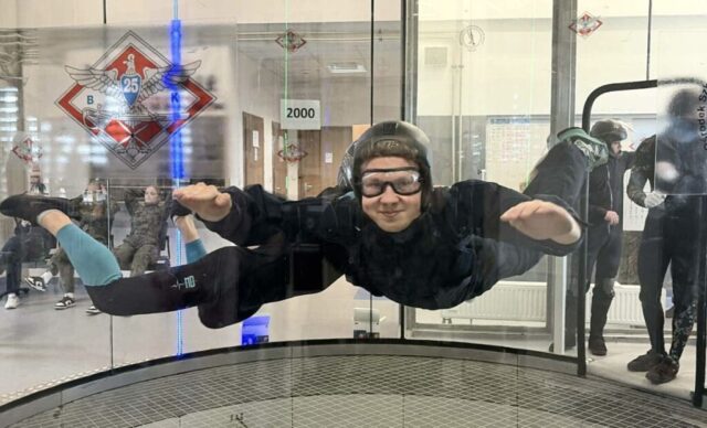 Osoba w tunelu aerodynamicznym utrzymuje pozycję w locie, przy obecności instruktora a inni obserwują zza szyby.