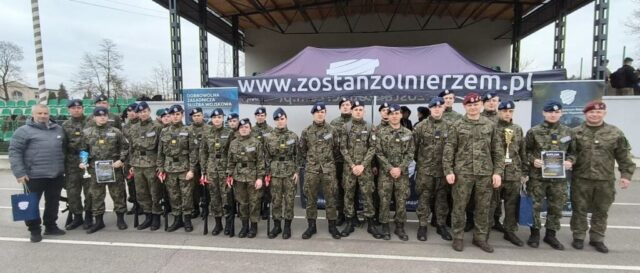 Grupa kadetów klasy OPW, żołnierzy w mundurach i jeden mężczyzna w stroju cywilnym stoi przed wojskowym namiotem rekrutacyjnym, trzymając dyplomy i trofea podczas uroczystości na otwartym terenie.