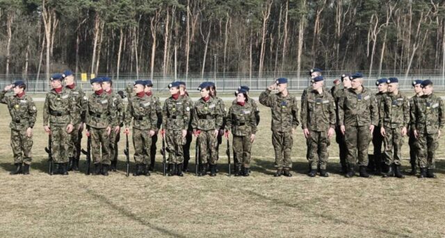 Pododdział kadetów klasy OPW w mundurach polowych i niebieskich beretach stoi w szyku na otwartym terenie, część z nich trzymając broń podczas oficjalnej ceremonii wojskowej