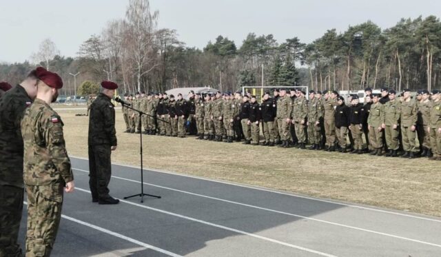 Żołnierz w bordowym berecie przemawia do mikrofonu podczas uroczystości wojskowej, a za nim stoją w szyku liczne pododdziały w różnych mundurach i nakryciach głowy.