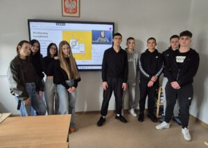 Grupa uczniów stoi w klasie przed ekranem z prezentacją o platformach handlowych.