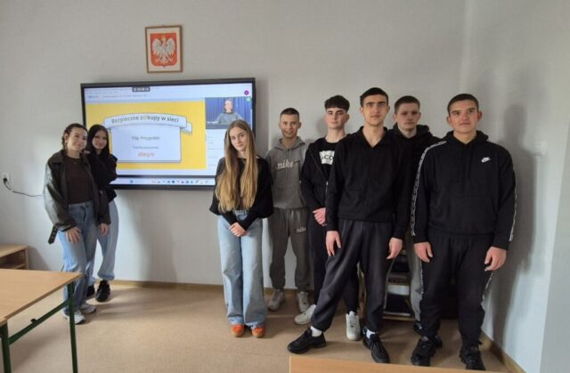 Grupa uczniów stoi w klasie obok ekranu z prezentacją o bezpiecznych zakupach online.