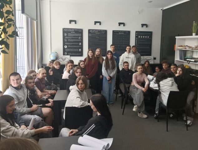 Duża grupa uczniów siedzących i stojących w sali konferencyjnej z plakatami edukacyjnymi i regałami z książkami.