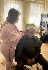 Mężczyzna siedzi na krześle a stojąca obok kobieta goli mu głowę maszynką.
