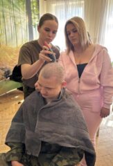 Uczeń klasy mundurowej siedzi na krzesełku. Za nim stoją dwie kobiety. Jedna goli mu głowę, a druga przygląda się temu.