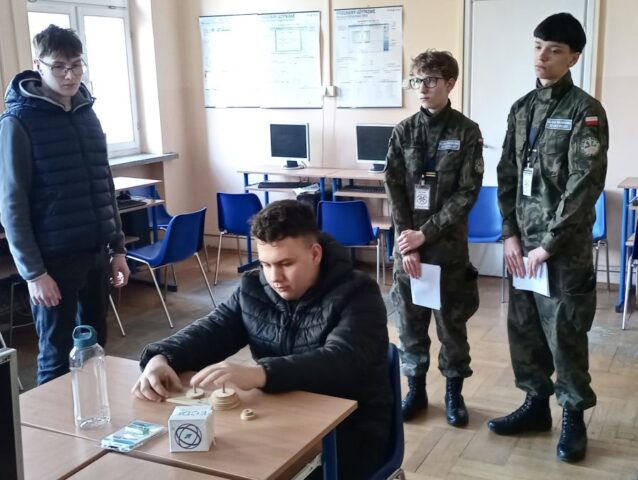 4 osoby w klasie informatycznej. Jedna z osób siedzi przy stoliku i układa wieże Hanoi.