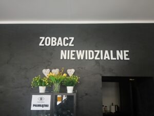 Ciemna ściana z napisem ‘ZOBACZ NIEWIDZIALNE’ oraz stojąca poniżej komoda z żółtymi tulipanami i dekoracjami.