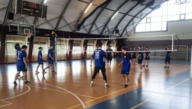 Dwie drużyny siatkówki rozgrywają mecz w hali sportowej; zawodnicy w niebieskich i ciemnych strojach stoją po obu stronach siatki, a sędzia obserwuje grę z podestu.