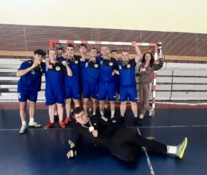 Szkolna drużyna piłki nożnej po zdobyciu medali pozuje wraz z opiekunem przed bramką w hali sportowej.”