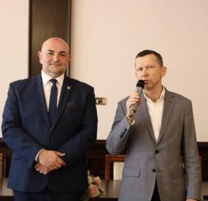 Dwóch mężczyzn w garniturach stoi w sali podczas uroczystości; jeden z nich przemawia do mikrofonu.