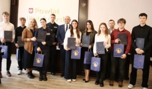 Grupa uczniów wraz z osobami dorosłymi pozuje podczas wręczenia stypendiów starosty; część trzyma teczki i torby z logo Powiatu Piotrkowskiego.