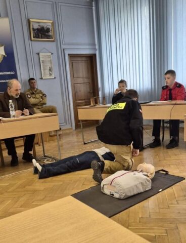 Uczeń klasy strażackiej demonstruje resuscytację na fantomie w sali szkoleniowej. W tle inni uczestnicy turnieju oraz organizatorzy.