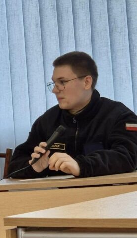 Osoba w mundurze z mikrofonem siedzi przy biurku podczas spotkania.