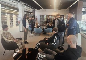 mediateka_wycieczka Grupa uczniów w bibliotece podczas oprowadzania. W tle regały z książkami.