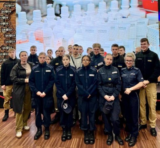 policyjne_targi5 Grupa uczniów w mundurach wraz z opiekunem i funkcjonariuszką policji pozuja do zdjęcia w sali szkoleniowej.