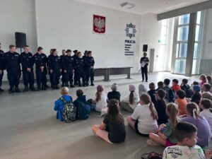 Kadeci klasy mundurowej stoją w szeregu podczas spotkania w sali, a policjantka prowadzi prelekcję przed grupą dzieci.