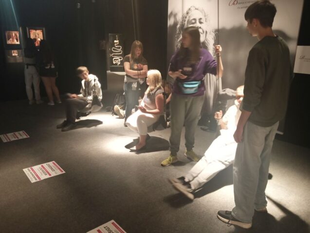 Grupa młodzieży wraz z opiekunem wewnątrz ciemnego pomieszczenia. W tle widoczne eksponaty wystawy body Worlds.