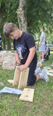 Uczestnik klęczy na trawie i łączy drewniane części w ramach zajęć ekologicznych.