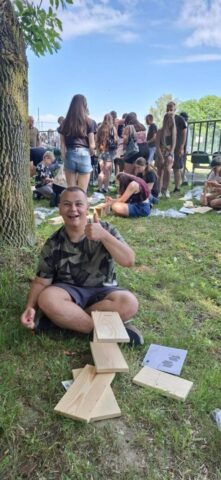 event_proekologiczny10 Osoba siedzi na trawie, pokazując gest aprobaty, otoczona drewnianymi elementami do montażu, w tle inni uczestnicy wydarzenia.