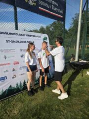 Igrzyska_LZS_final_centralny1 Trzy osoby stoją na trawie podczas dekoracji medalowej przy banerze wydarzenia sportowego.