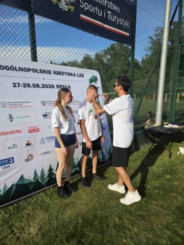 Igrzyska_LZS_final_centralny1 Trzy osoby stoją na trawie podczas dekoracji medalowej przy banerze wydarzenia sportowego.