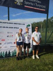 Igrzyska_LZS_final_centralny3 Trzy osoby w strojach sportowych stoją na trawie przed banerem wydarzenia LZS.