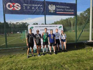 Igrzyska_LZS_final_centralny4 Sześć osób w strojach sportowych stoi na boisku przed banerem ogólnopolskich igrzysk LZS.