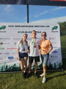 LZS_final_centralny Dwoje uczniów z medalami wraz z opiekunem podczas zawodów sportowych. W tle baner imprezy pod nazwą XXIX Ogólnopolskie Igrzyska LZS w Spale.