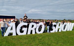 Grupa uczniów wraz z opiekunem pozująca przy dużym napisie „AGRO SHOW” na terenie wydarzenia rolniczego.