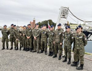 Grupa uczniów w mundurach uczestniczy w wyjeździe edukacyjnym, stojąc przed jednostką wojskową.