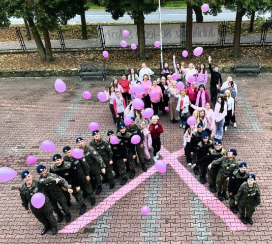 Uczniowie ustawieni na dziedzińcu wokół symbolu różowej wstążki, trzymający różowe balony.