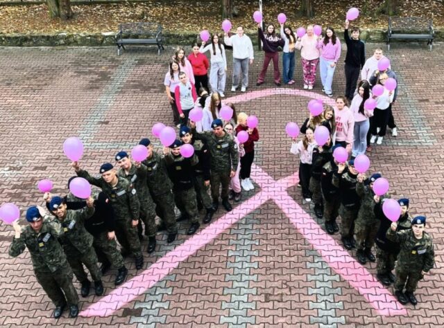 Uczniowie i nauczyciele z różowymi balonami wokół symbolu różowej wstążki na dziedzińcu szkoły.