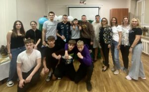 Grupa uczniów i nauczycieli podczas wspólnego zdjęcia w sali. W tle ekran projekcyjny i dekoracje z balonów.