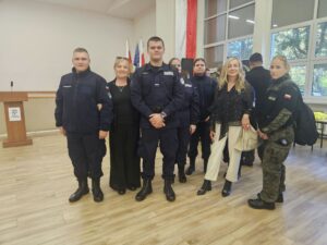 Grupa uczniów w umundurowaniu wraz z nauczycielami pozuje do zdjęcia w sali podczas wydarzenia. W tle widoczne są flagi oraz uczestnicy spotkania.