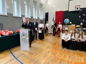 Uroczystość szkolna w sali gimnastycznej. Osoba przemawia przy mównicy, obok stoją uczniowie oraz poczet sztandarowy na tle biało-czerwonej dekoracji.