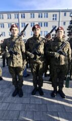 Trzy osoby w mundurach wojskowych stojące przed budynkiem podczas uroczystości wojskowej.