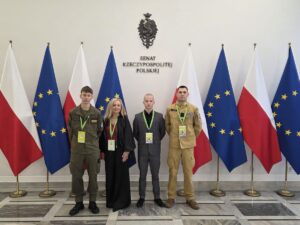 wycieczka_sejm 3 uczniów wraz z opiekunem stoją przed flagami Polski i Unii Europejskiej w budynku Senatu Rzeczypospolitej Polskiej.